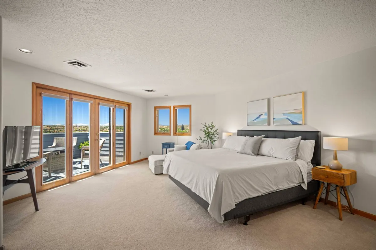 A serene bedroom setup featuring a large bed with white linens, sliding glass doors that open to the outdoors, decorative artwork on the wall, and a nightstand with a lamp. Natural light fills the space, highlighting its calming ambiance and strong connection to the surrounding landscape.