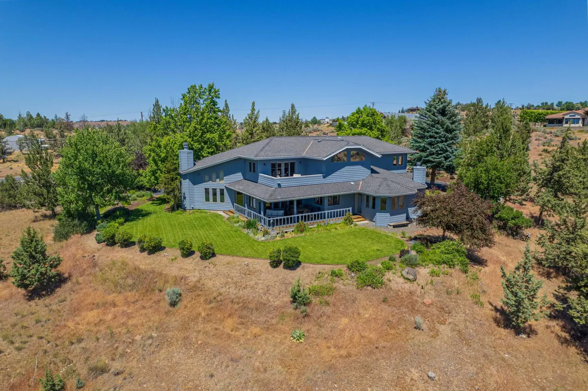 The image showcases a charming single-family home perched on a manicured hillside lawn, surrounded by mature trees. The inviting porch offers sweeping views of the sunset over the valley, while the spacious yard hints at space for outdoor gatherings and relaxation in nature.