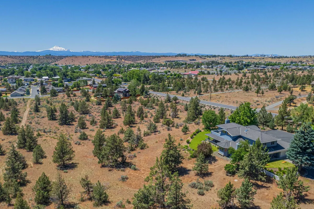 A sweeping view of distant snow-capped mountains, a sprawling residential area with greenery, and a winding road leading toward outdoor adventure. The vast open sky and tranquil setting evoke a sense of peace and connection to nature.