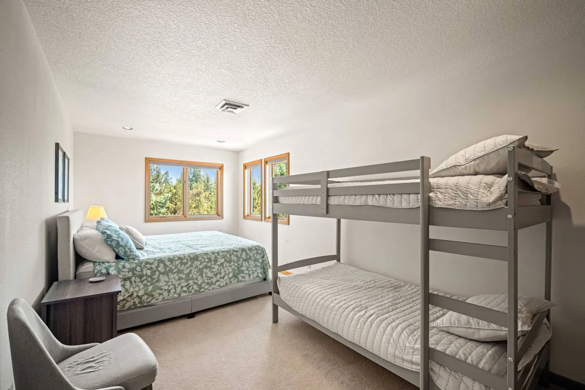 A cozy family-friendly bedroom featuring a queen bed and a bunk bed, with windows that let in morning sunlight, a nightstand with a lamp, and a relaxed, inviting mood. The setup is ideal for parents and children sharing one space.