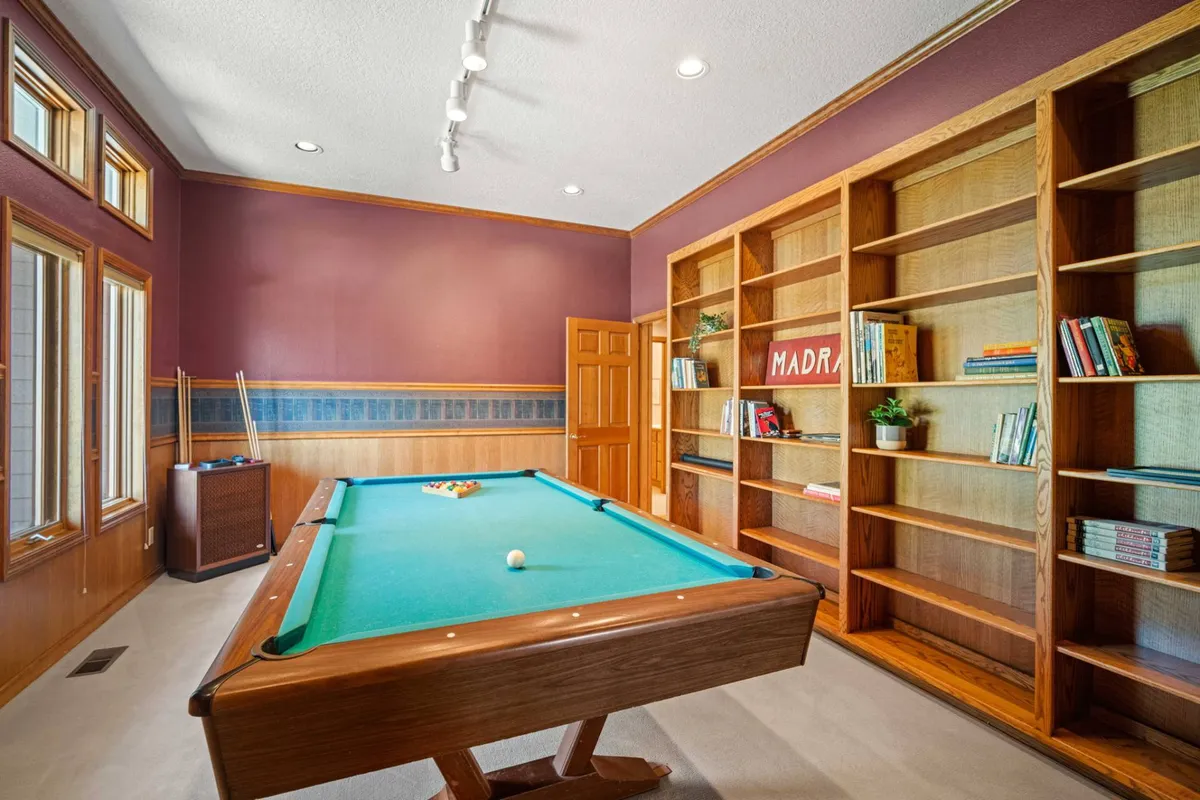 A relaxed and inviting game room featuring a pool table as the centerpiece, flanked by bookshelves and large windows that fill the space with natural light. The setup encourages both spirited billiards matches and casual socializing, with cozy touches that foster a sense of togetherness.