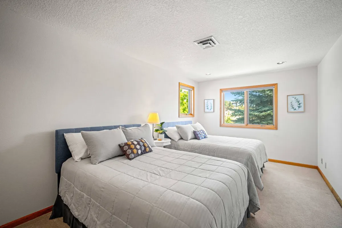 A calm and cozy bedroom with two beds adorned with decorative pillows, a lamp on the side table, and windows that look out onto greenery. The space is inviting for relaxation, reading, or sharing quiet moments.