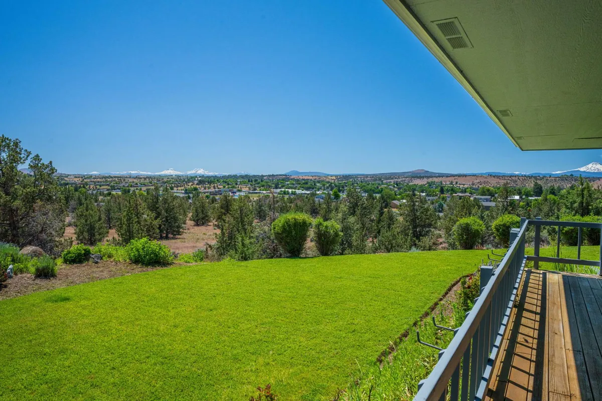 This view captures a sweeping mountain range beyond an expansive green lawn, with the wooden deck's railing in the foreground. The scene evokes a connection to Oregon’s high desert beauty and highlights the tranquil, scenic environment for relaxation or entertaining outdoors.