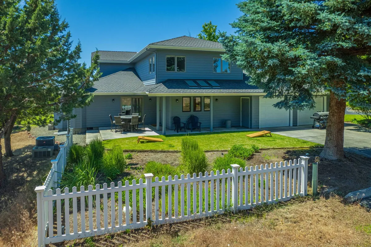 A welcoming view of the home's exterior highlights a green lawn bordered by a white picket fence, with a deck equipped with comfortable seating and large windows connecting the inside and outside. The inviting setup showcases both the deck and yard, offering direct access for relaxation or pet play.