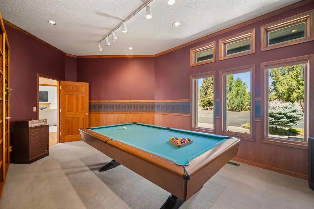 A playful, sunlit interior where a pool table takes center stage, surrounded by large windows and warm wooden cabinetry. The space suggests lively game nights and easy camaraderie among guests.