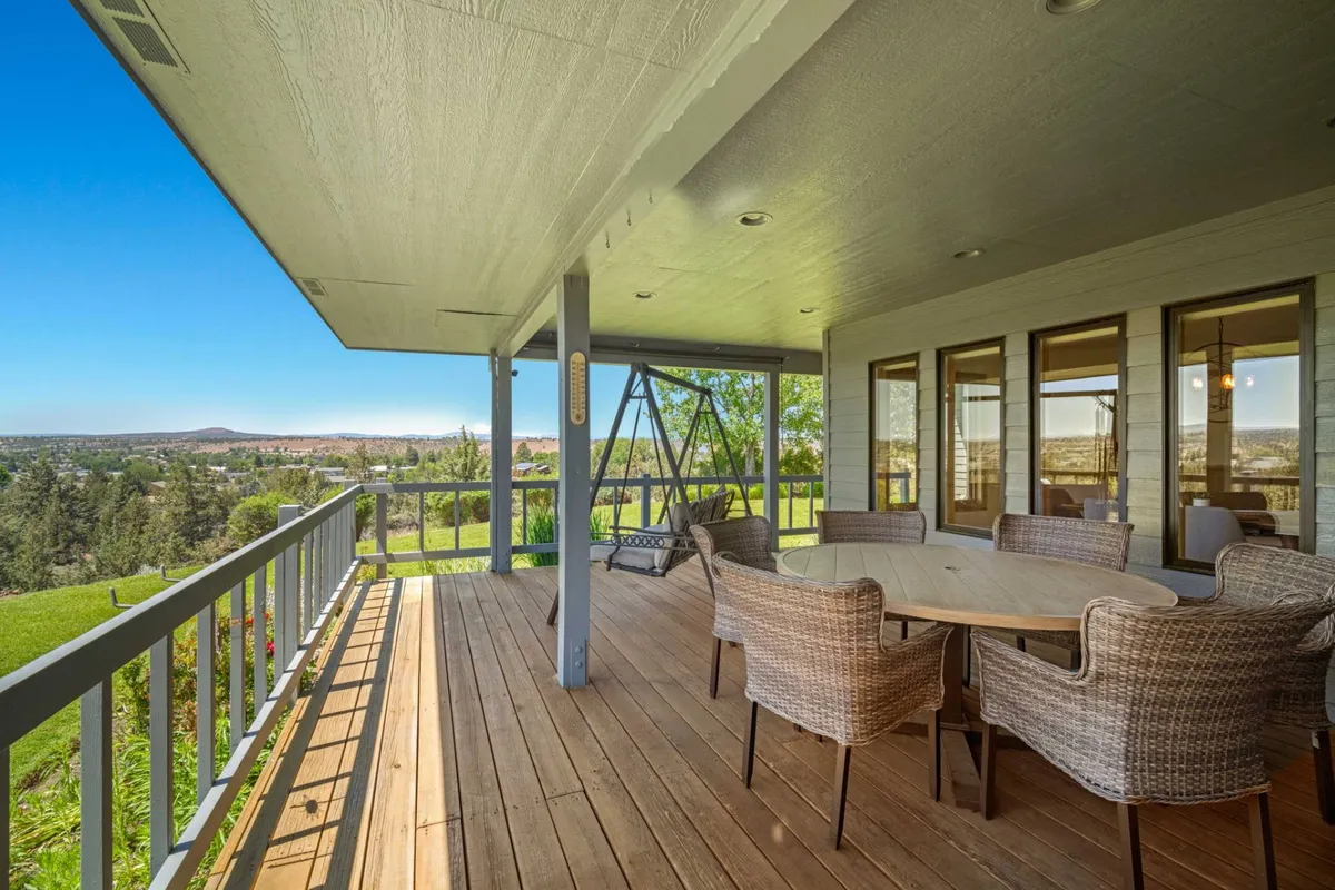 A spacious outdoor deck features a dining table with wicker chairs and a comfortable swing. The scene is inviting, with ample seating for a group to gather for meals, drinks, or games. The backdrop includes lush greenery and an open view, perfect for evening gatherings or morning relaxation.